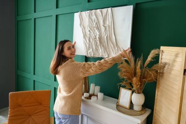 Genç bir kadın evdeki şöminenin üstündeki yeşil duvara resim asıyor.