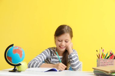 Küçük kız sarı arka plandaki masada ders alıyor.