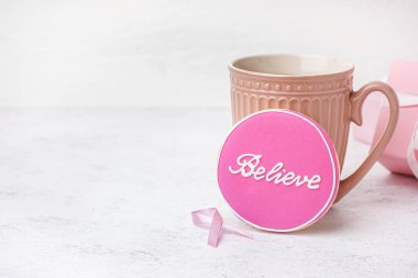 BELIEVE yazılı pembe kurabiye, beyaz grunge arka planda bir fincan çay ve kurdele. Göğüs kanseri farkındalığı konsepti