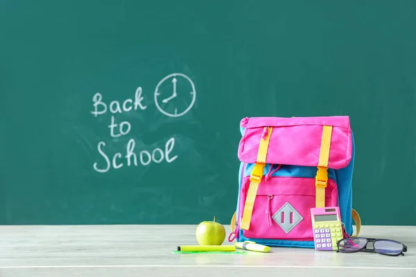 Backpack and supplies on table near green chalkboard with text BACK TO SCHOOL
