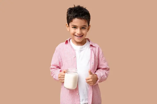 Cute little boy with mason jar of milk showing thumb-up on beige background
