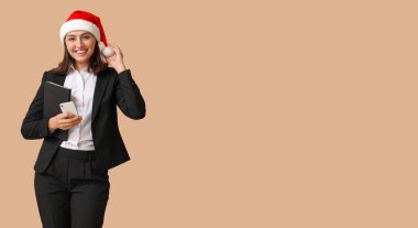 Noel Baba şapkalı güzel bir iş kadını. Not defteri ve cep telefonu bej arka planda. Mesaj için yeri var.