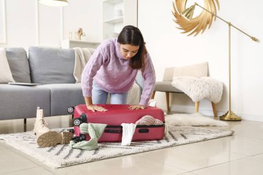 Evde bir sürü kıyafetle valizi kapatmaya çalışan genç bir kadın.