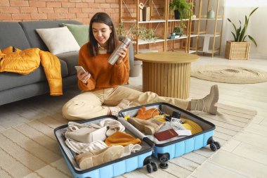 Cep telefonu ve termosu olan genç bir kadın evde kışlık giysilerini bavula koyuyor.