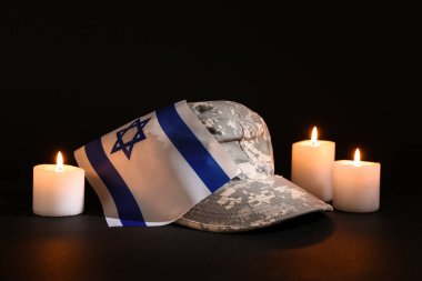 İsrail bayrağı taşıyan askeri şapka ve koyu arkaplanda yanan mumlar.