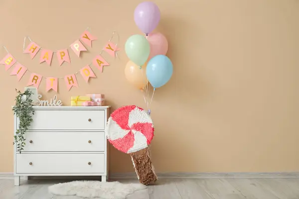Chest of drawers, pinata, balloons and garland near beige wall in living room decorated for birthday