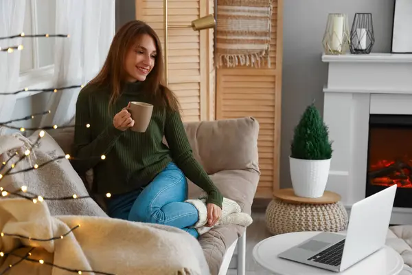 Young woman with cup of cocoa watching video at home