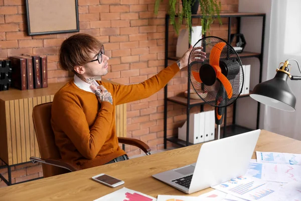 Sweaty young man with electric fan in office