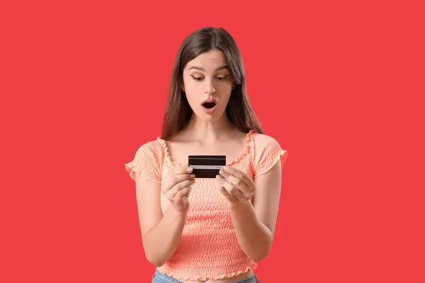 Shocked young woman with credit card on red background
