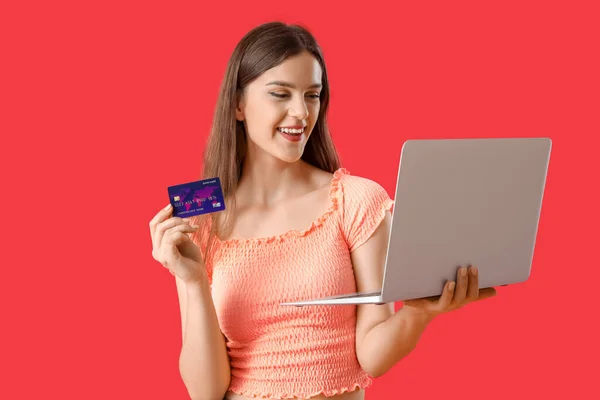 Young woman with credit card and laptop on red background
