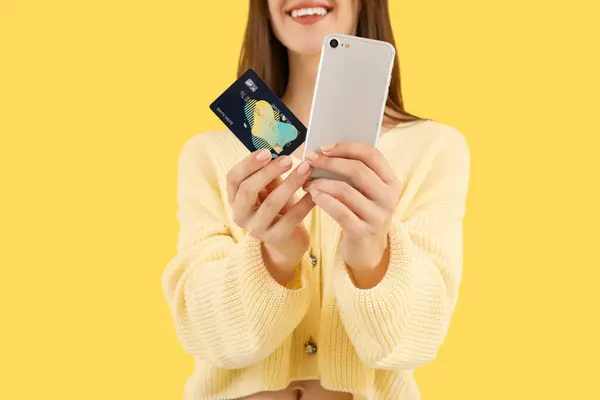 Young woman with credit card and mobile phone on yellow background, closeup