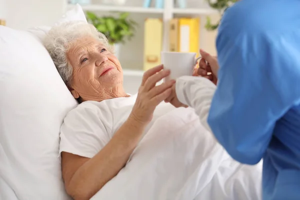 stock image Senior woman taking cup of tea from nurse in bedroom