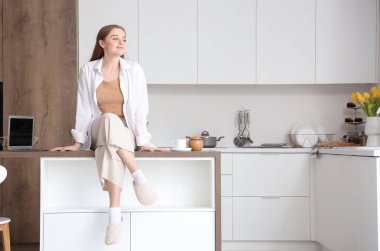 Masada oturan genç ve güzel bir kadın modern dizüstü bilgisayarla aydınlık mutfakta.