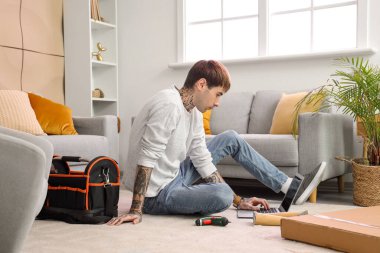 Genç adam evde mobilya montajı yaparken dizüstü bilgisayar kullanıyor