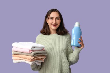 Young woman with clean towels and bottle of detergent on lilac background clipart