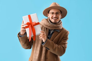 Mavi arka planda Noel hediyesi olan kışlık elbiseli genç adam.