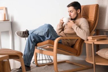 Evdeki koltukta oturan bir fincan sıcak çayla genç adam.