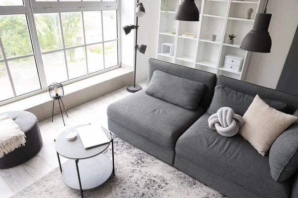 Interior of light living room with cozy grey sofa and coffee table near big window