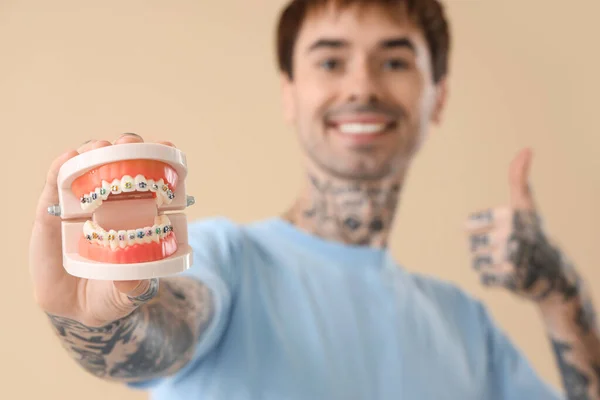 Young tattooed man holding jaw model with braces on beige background, closeup