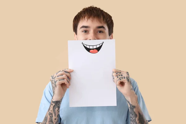 Young tattooed man with paper smile on beige background