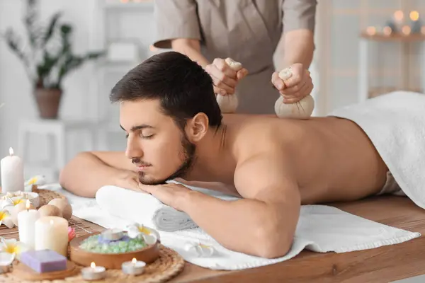 Young man undergoing treatment with herbal bags in spa salon, closeup