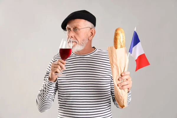 Senior man with flag of France and baguette drinking wine on light background