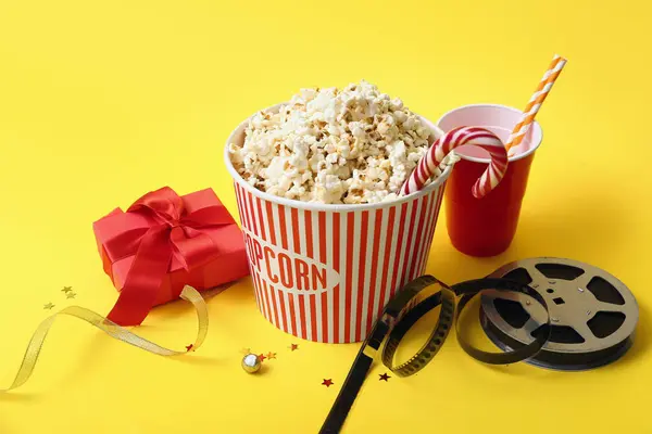 Bucket of popcorn with drink, film reel and Christmas gift on yellow background