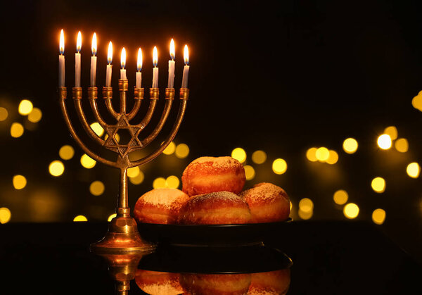 Менора с горящими свечами и пончиками для празднования Хануки на темном столе против размытых огней