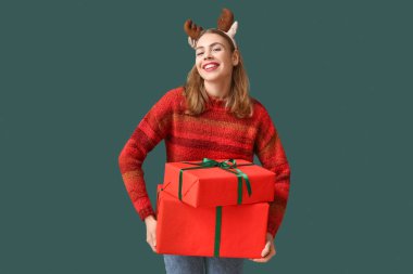 Geyik boynuzları takmış, yeşil arka planda Noel hediyesi kutuları olan güzel genç bir kadın.