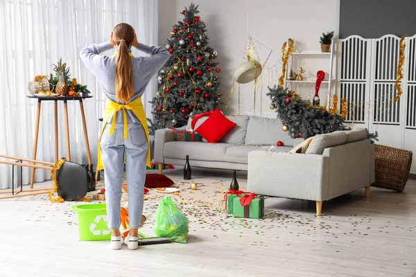 Shocked female janitor in messy living room after New Year party, back view