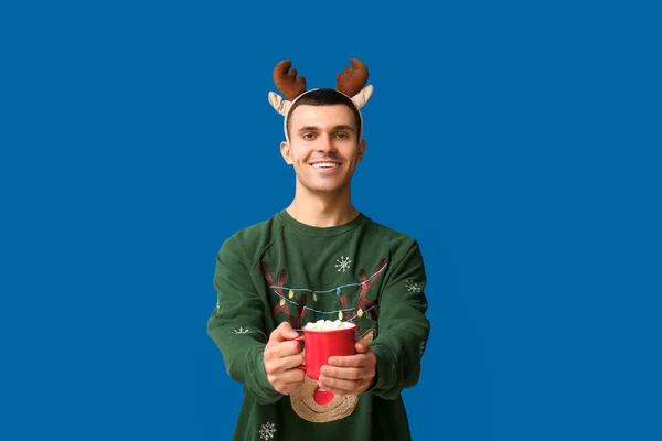 stock image Young man in reindeer horns holding cup of coffee with marshmallows on blue background