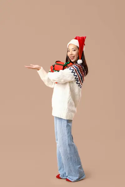 茶色の背景にクリスマスギフトボックスとサンタの帽子の若いアジアの女性 — ストック写真