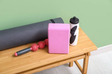 Yoga minderli ahşap masa, blok, masaj silindiri ve yeşil duvarın yanında bir şişe su.