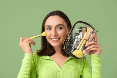 Makyaj ürünleriyle dolu kozmetik çantasıyla ve yeşil arka planda fırçasıyla güzel bir genç kadın.