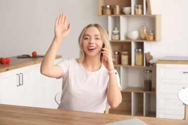 Güzel genç bir kadın cep telefonuyla konuşuyor ve evde el sallıyor.