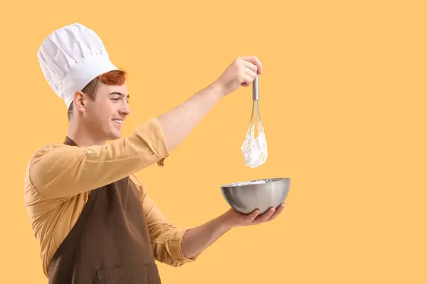Male confectioner in apron with bowl and whisk preparing tasty meringue on yellow background