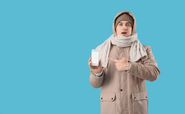 Kışın giyinmiş genç adam mavi arka planda cep telefonuna bakıyor.