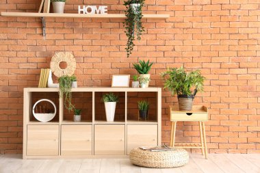 Farklı ev bitkileri, kitaplar ve kahverengi tuğla duvarın yanında boş fotoğraf çerçevesi olan raf ünitesi.