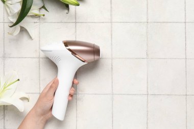 Beyaz karo arka planda modern fotoepilatör ve zambak çiçeklerine sahip kadın eli.