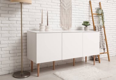 Odada beyaz tuğla duvarın yanında merdiveni ve lambası olan modern beyaz çekmeceler.