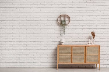 Modern hafif ahşap çekmeceler içinde lamba ve vazoda beyaz tuğla duvarın yanında otlar var.