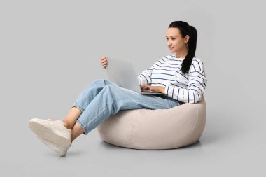 Dizüstü bilgisayarı olan güzel genç bir kadın gri arka planda rahat bir puf koltukta oturuyor.