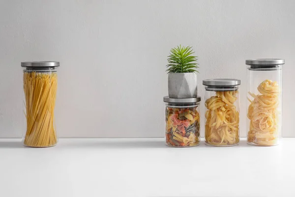 Glass jars with different types of pasta and houseplant on white kitchen counter