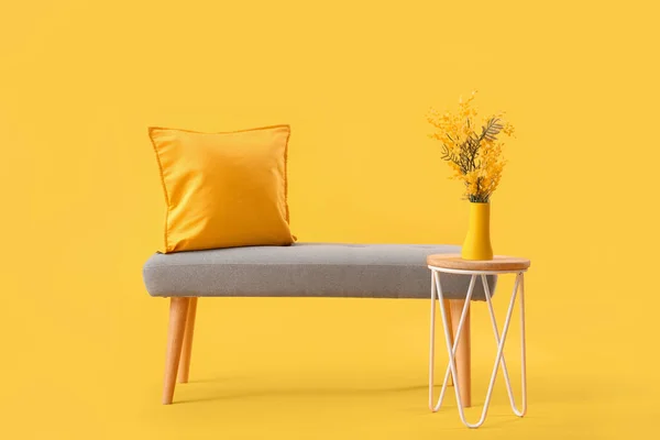 Grey bench with cushions and flowers on coffee table against yellow background