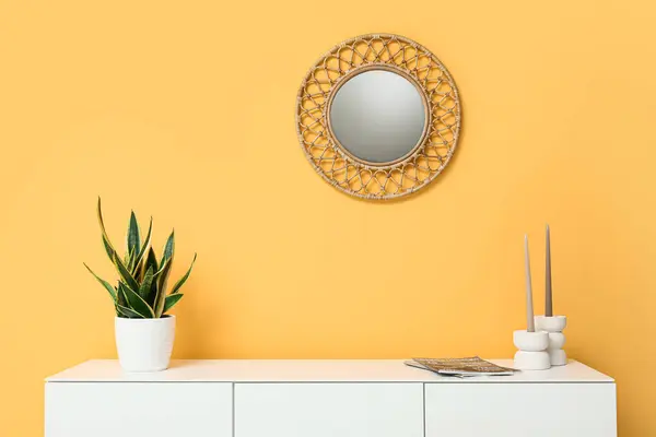 Modern white chest of drawers with houseplant and mirror near orange wall in room