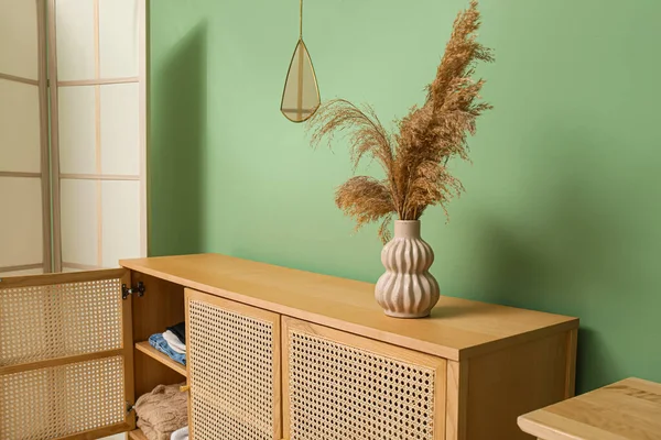 Modern light wooden chest of drawers with clothes and pampas grass in vase near green wall in room