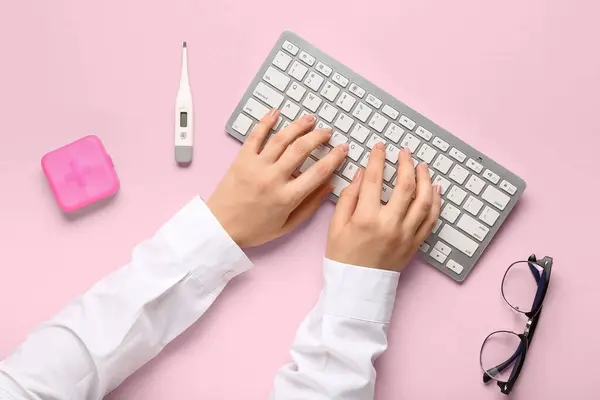 Doctor working with computer keyboard on pink background, top view