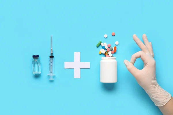 Doctor showing OK with pills, paper cross, syringe and vaccine on blue background