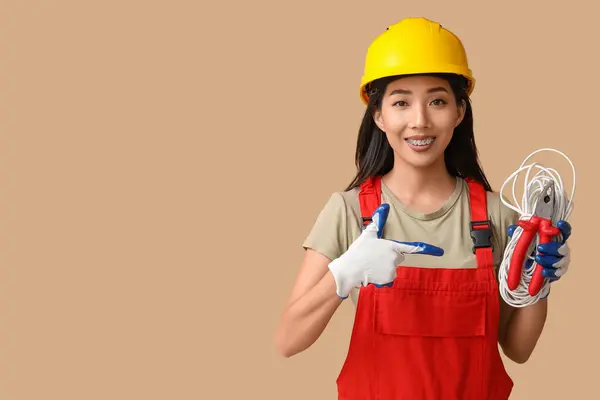 Young female electrician pointing at pliers and cables on beige background