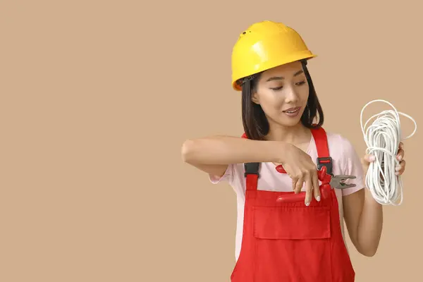 Young female electrician with pliers and cables on beige background